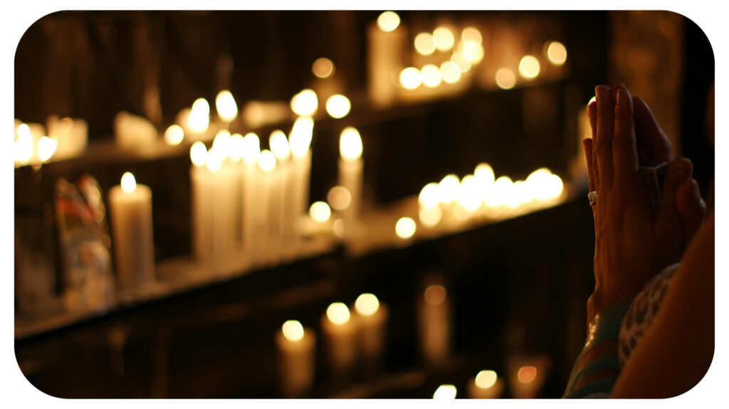 A person prays with clasped hands before rows of glowing candles, creating a serene and spiritual atmosphere.
