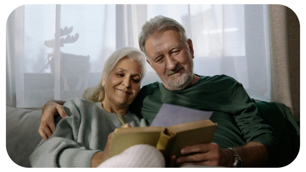 An elderly couple sits close together on a couch, smiling warmly as they read a book and a letter, reflecting love, comfort, and cherished memories.