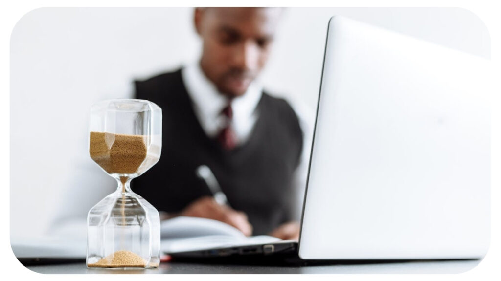 A clear hourglass with golden sand stands in focus, symbolizing time, as a man works on his laptop and writes in a notebook in the background.