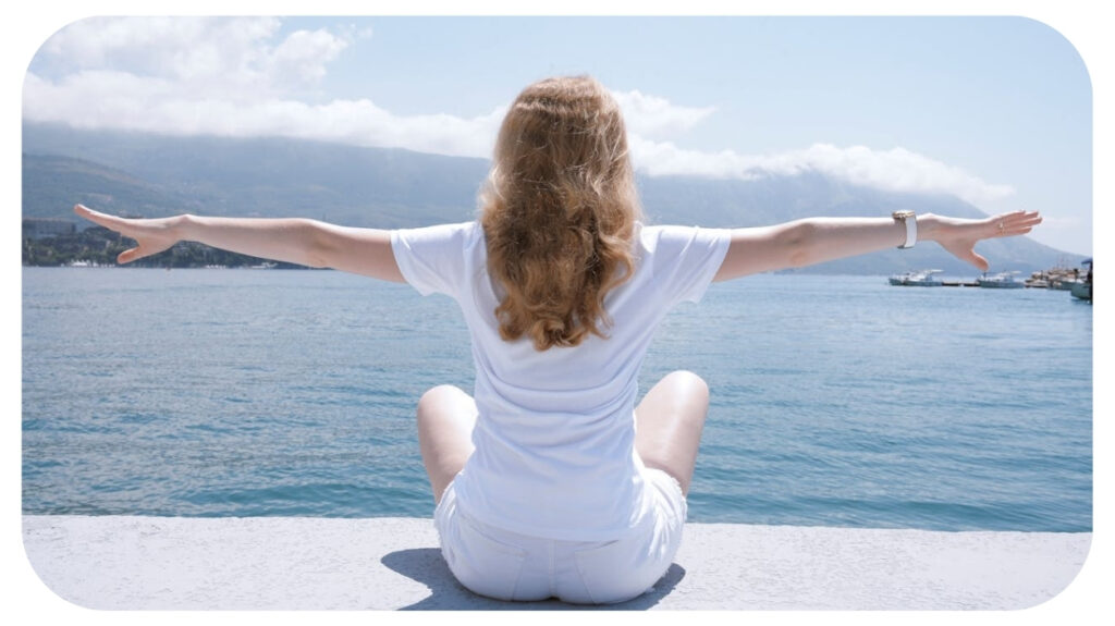A woman in white sits by the sea with arms wide open, embracing the serene view of mountains and water.