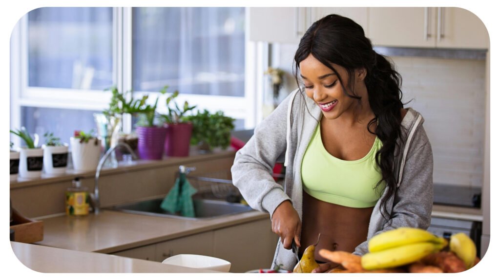 A cheerful woman in activewear slices a pear in a bright kitchen, surrounded by fresh fruits and plants, reflecting a healthy lifestyle.