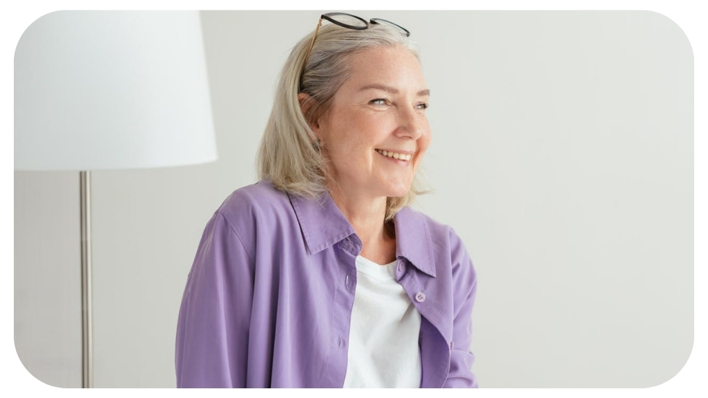 Smiling senior woman with gray hair wearing a purple shirt and white top, sitting indoors with eyeglasses on her head and a lamp in the background.