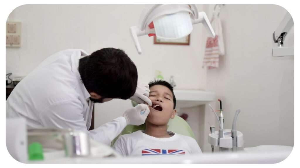 A young boy receiving a dental check-up from a pediatric dentist, mouth open under an overhead examination light in a clinic.