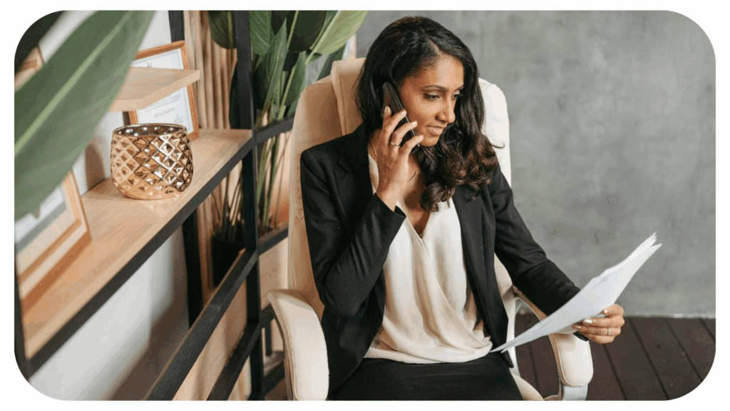 A professional woman sitting in an office chair, talking on the phone while reviewing documents, with a decorative shelf and plants in the background.