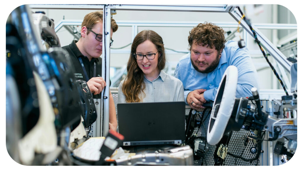 Engineers working together on an advanced manufacturing system, analysing data on a laptop surrounded by connected machines and automotive components, representing the concept of an integrated factory where machines and data communicate for seamless production.