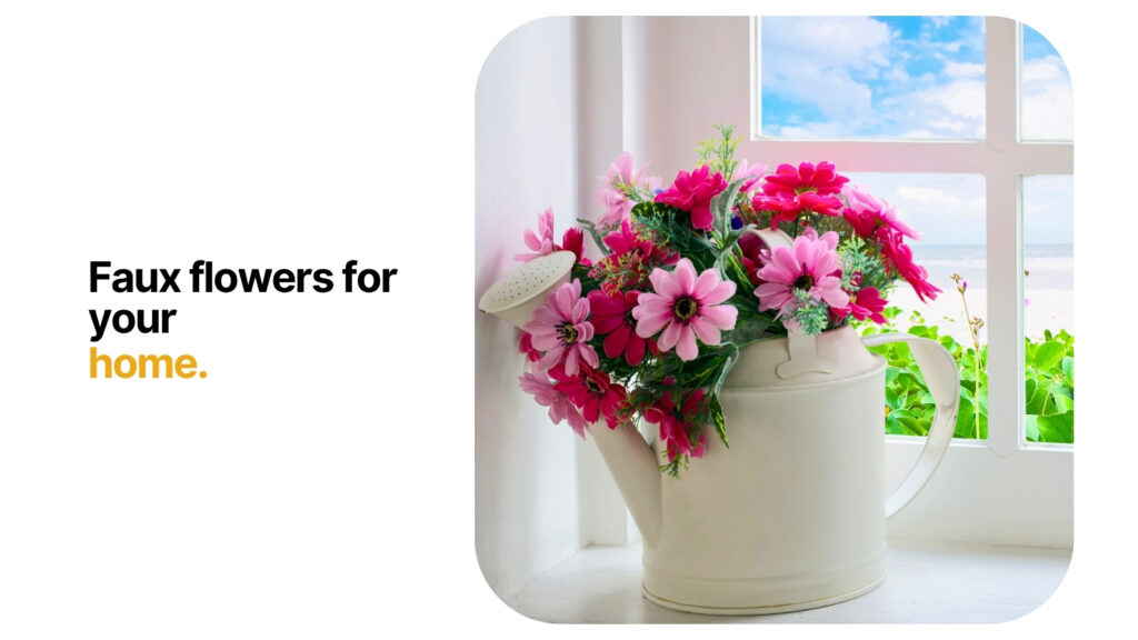 A white watering can filled with bright pink faux flowers sits on a windowsill, with sunlight streaming through a window overlooking a scenic outdoor view, creating a cheerful and decorative home atmosphere.