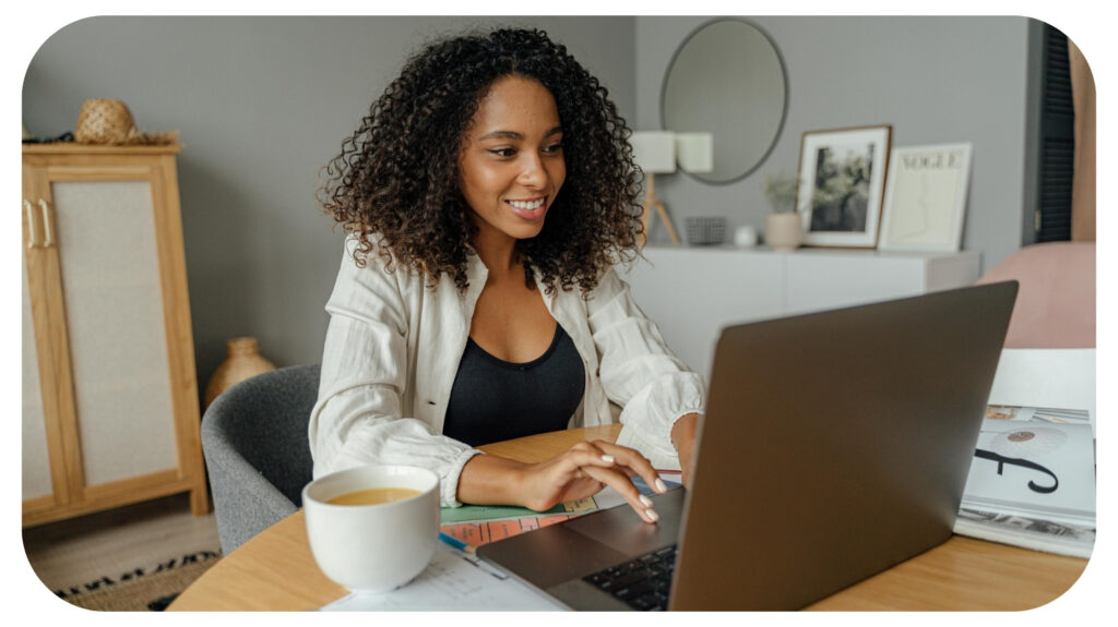A young woman smiling while working on her laptop at a home desk with a cup of coffee, papers, and magazines around, symbolising focus, balance, and the modern remote work lifestyle featured in the Most Inside article.