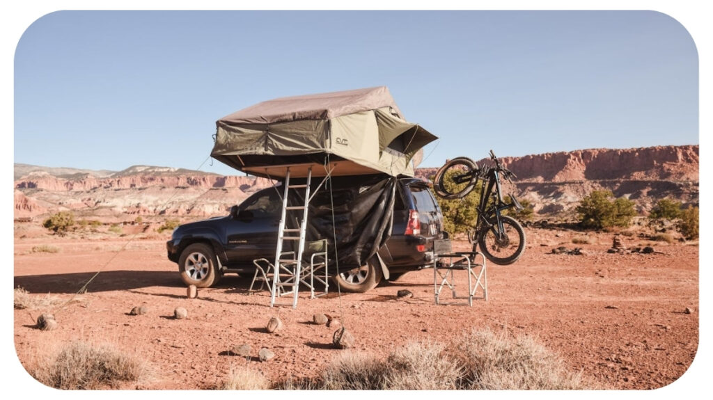 SUV with rooftop tent camping setup parked in desert landscape with ladder chairs and bicycle mounted for overland travel adventure.