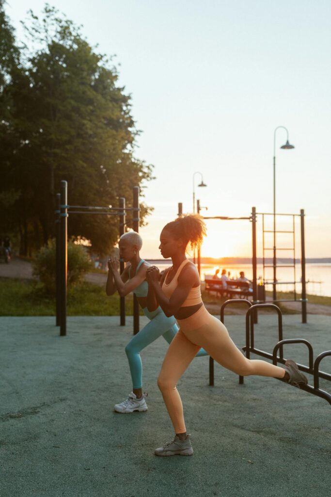 two women performing outdoor strength training exercises wearing supportive athletic wear and workout leggings during a sunset fitness session at a public workout park.